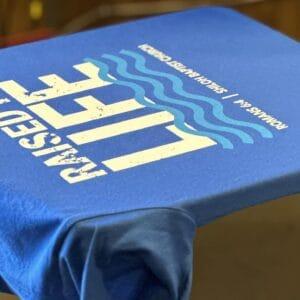 A royal blue church tee screen printed with a Raised Life design and wave motif