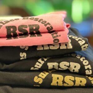 A folded stack of RSR Surface Restoration tees in pink, black and grey