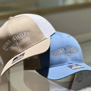 Two Selma Jewelry trucker caps, one tan and one blue, with embroidered logos
