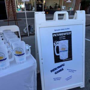 Printed Oktoberfest beer steins stacked on a table beside the sign advertising them