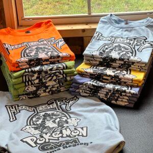 Stacks of folded Hidden Temple Toys tees in six colours, each carrying the same printed badge