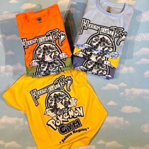 Folded Hidden Temple Toys tees in orange, white and yellow against a cloud backdrop