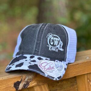 A CW Ranch trucker cap resting on a wooden rail, showing the embroidered logo and cow print brim