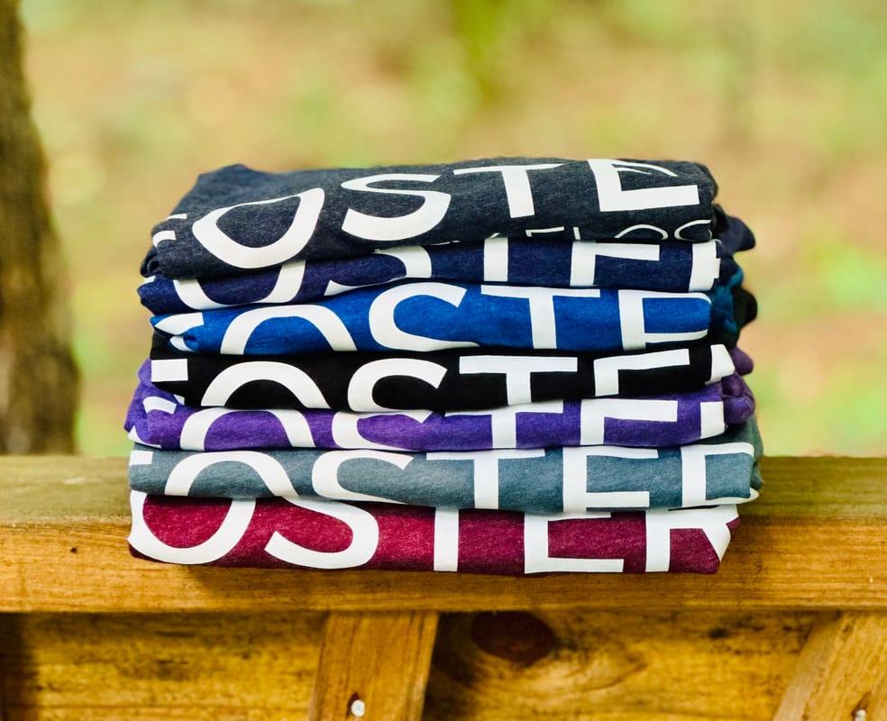 A stack of folded Foster tees in seven heathered colours, resting on a wooden rail