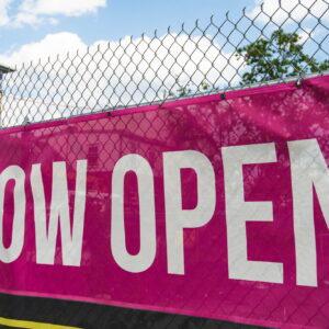 A pink Now Open vinyl banner fixed to a chain link fence